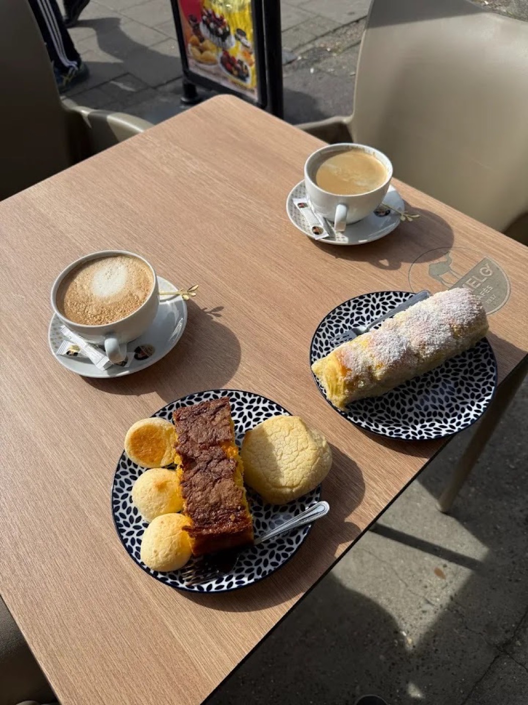 Two coffees with pão de queijo, a slice of cake and a sugar-dusted roll on a sunny table